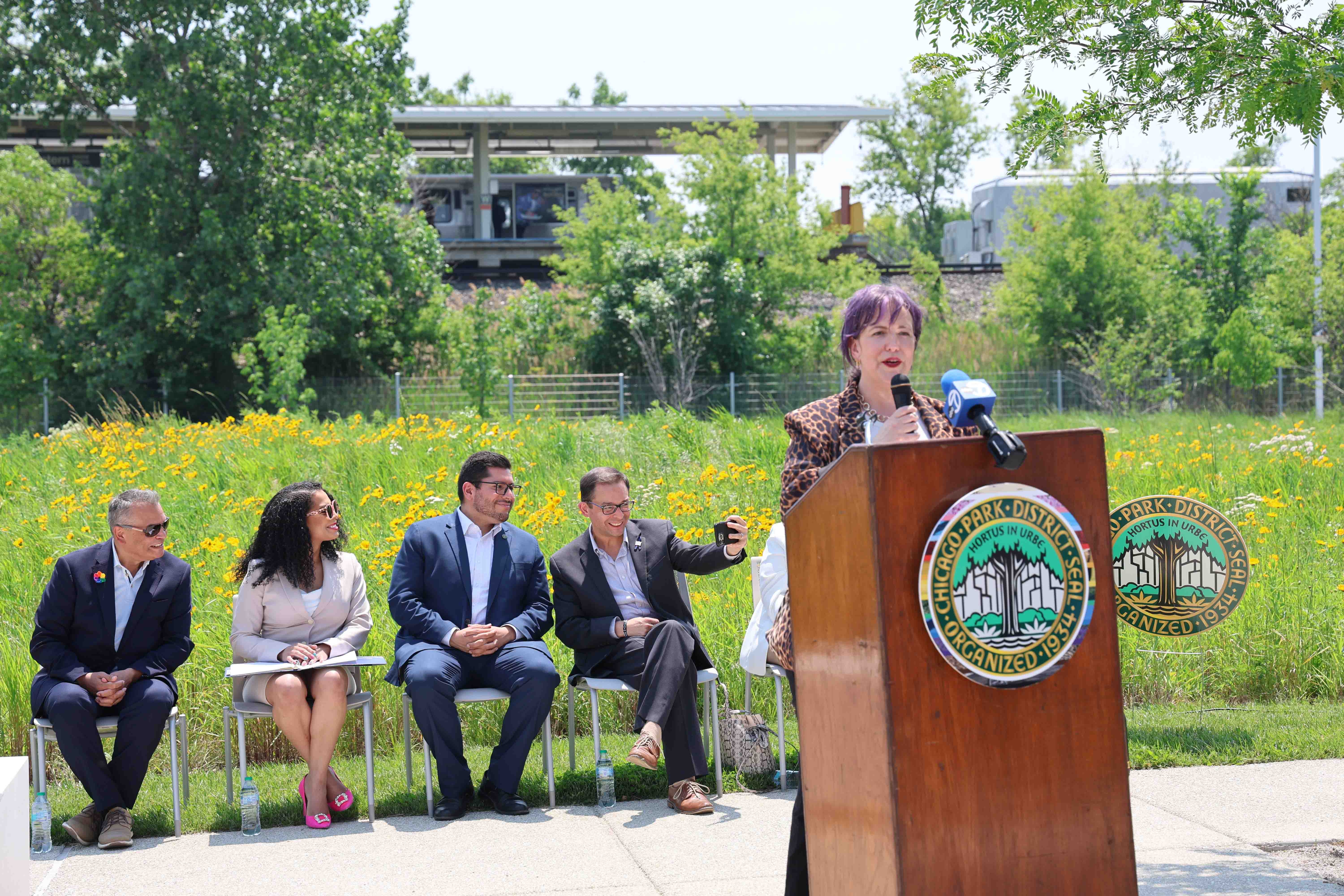 Woman speaks at podium with officials seated nearby, train station and wildflowers in the background.
